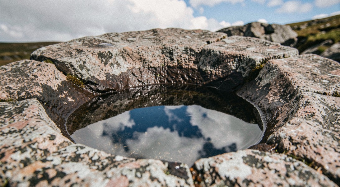 A shallow rock basin scoured into the top of a Dartmoor granite tor, still rainwater in the bowl reflecting a blue sky and cloud, coarse crystalline granite around it, close overhead framing