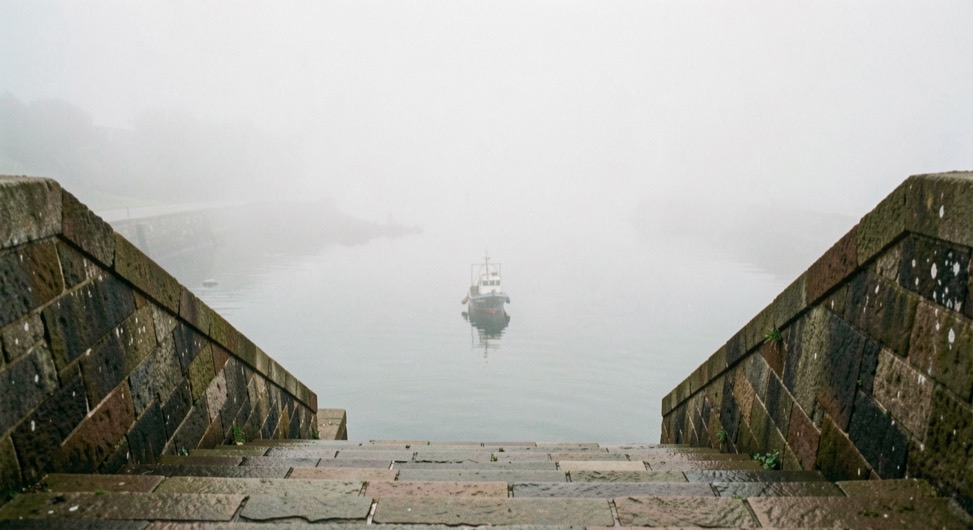 Stone harbour steps descending into still water in dense sea fog, a small fishing boat in silhouette a few metres away, the far side of the harbour invisible in white, soft even light, upright framing