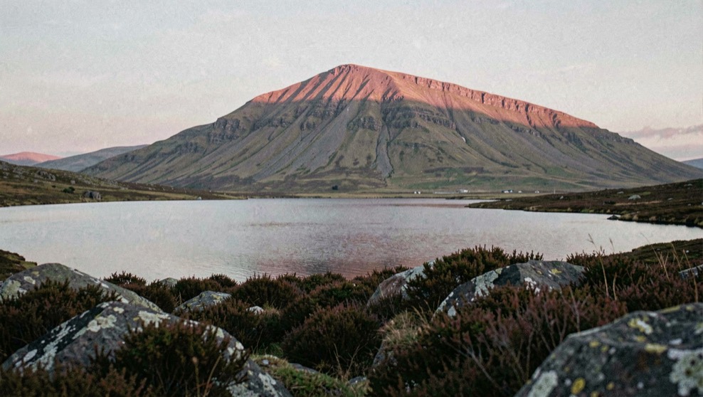 A still Highland loch at dawn with an isolated steep sided mountain rising behind it, dark heather and rock in the foreground, cold pink light along the summit ridge, wide horizontal framing
