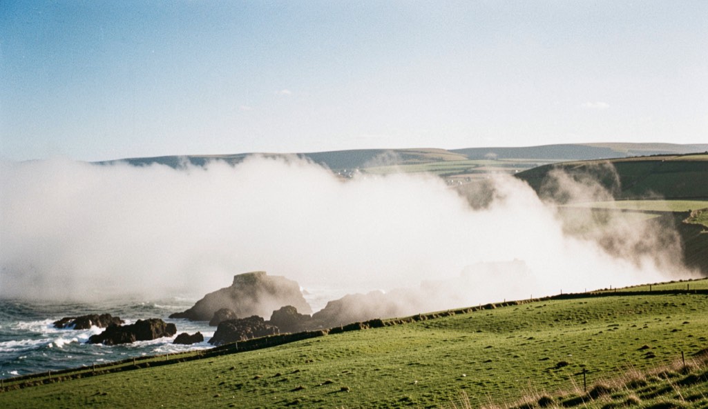 A dense white bank of sea fog lying just offshore along the Fife coastline in the afternoon, clear blue sky above the land, dark rocks and green fields in sunlight in the foreground, wide horizontal framing