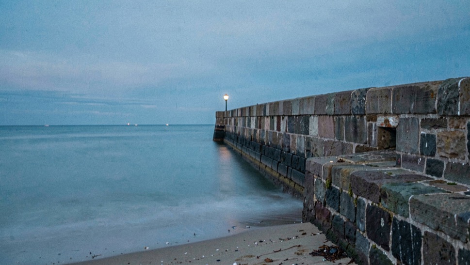 A stone harbour arm running out into a smoothed sea during a long exposure at dusk, a single lamp lit at the far end, blue light in the sky above the horizon, wide horizontal framing from the beach beside the wall