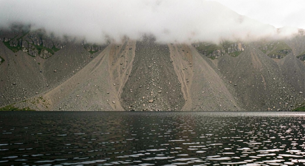 Black still water in the foreground with a long wall of loose scree falling directly into the lake behind it, low cloud along the ridge above, thin light from the right, wide horizontal framing