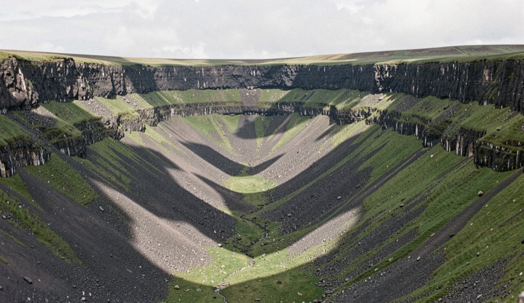 A deep symmetrical U shaped valley cut into the north Pennine escarpment, a band of dark dolerite rock running along both rims, grass and scree below, cloud shadow crossing the valley floor, wide horizontal framing