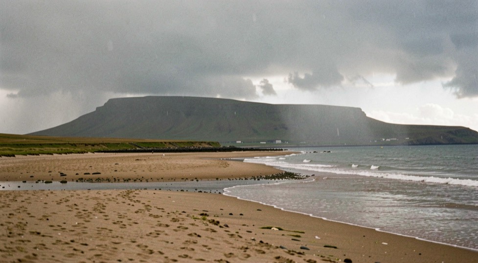 A rain shower crossing the Solway Firth seen from the Cumbrian sand flats, a grey curtain of rain reaching the water on the right, the hill of Criffel dark against a lit sky on the far shore, wide horizontal framing