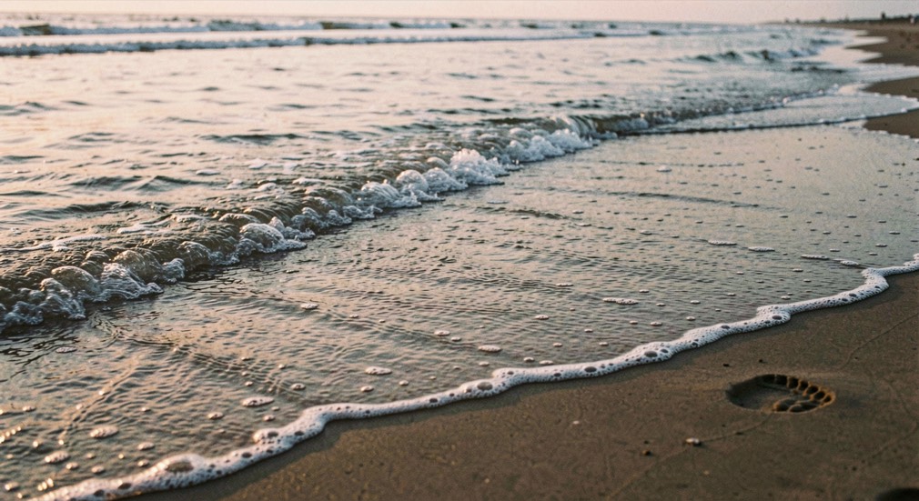 A shallow sheet of returning tide water running fast across flat sand at evening, the leading edge catching low light, footprints being erased ahead of it, wide low framing along the sand